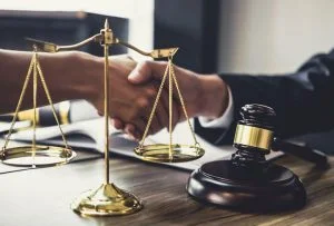 A golden scale of justice and a wooden legal gavel on a lawyer's desk, with two professionals shaking hands in the background to seal a legal agreement.